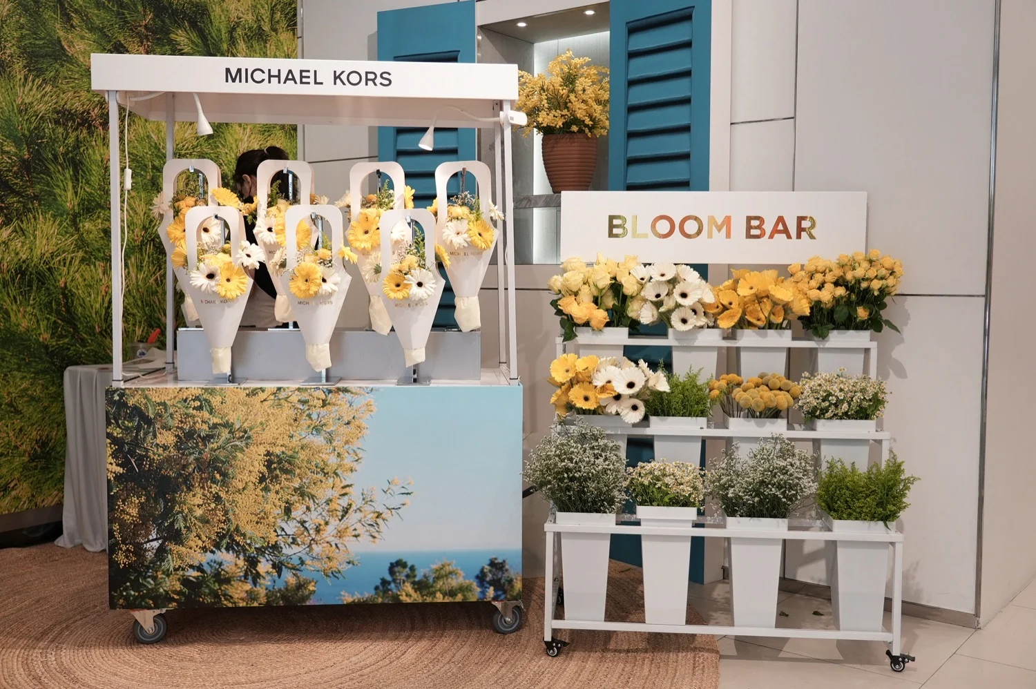 Michael Kors Bloom Bar floral cart with yellow and white flowers beside a blue-shutter backdrop