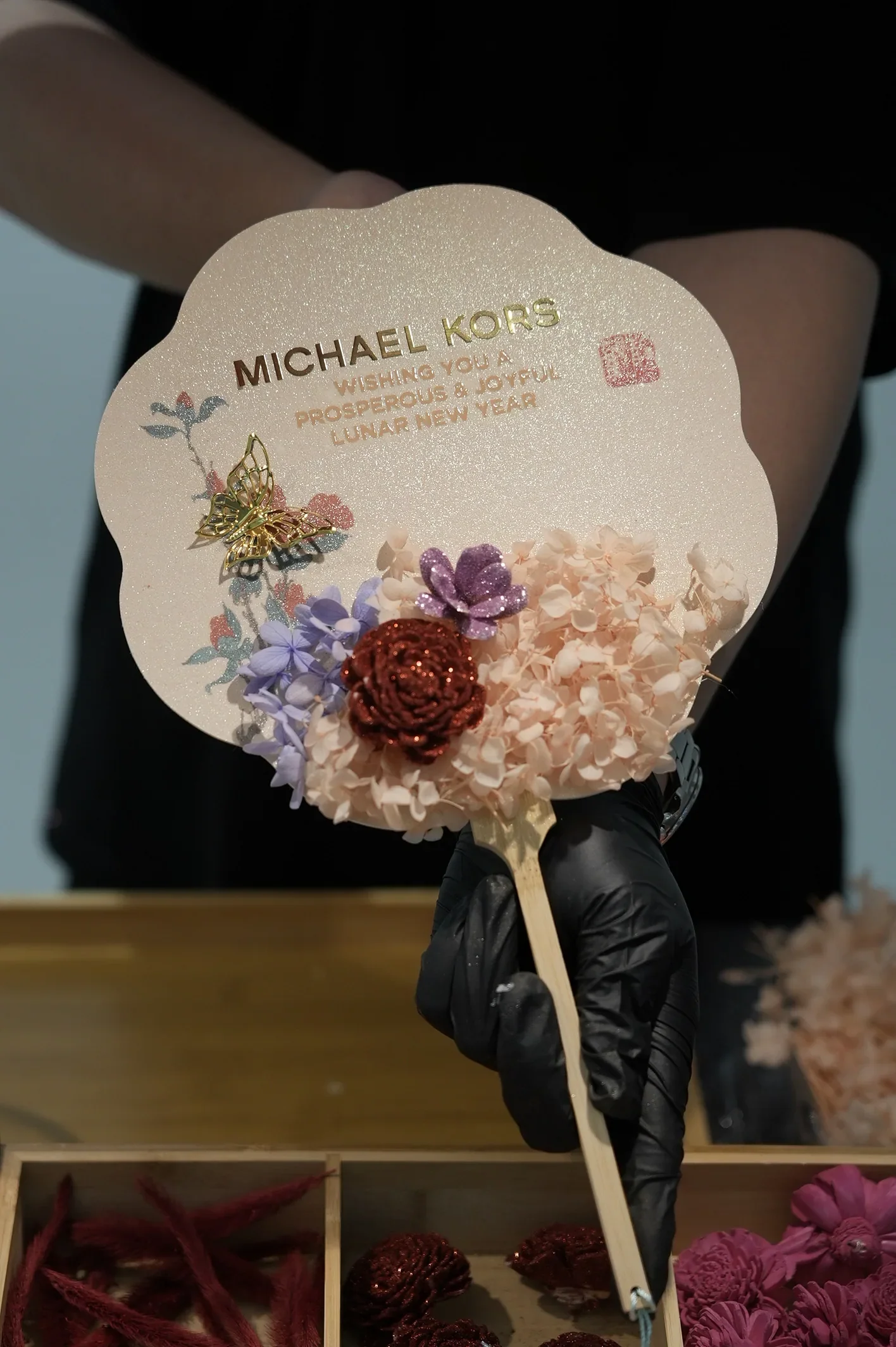 Gloved hands presenting a Michael Kors fan decorated with small purple and pink flowers